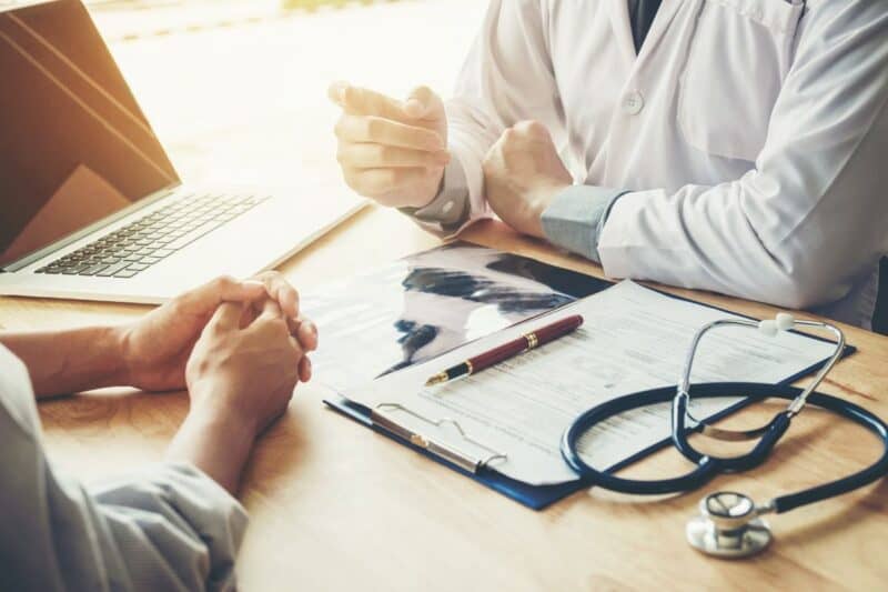 Healthcare professional talking to a patient with an x-ray, clipboard, stethoscope and computer on the desk