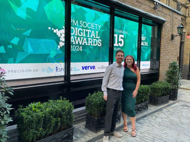 Ellie and Craig stand outside signage for The PM Society Digital Awards